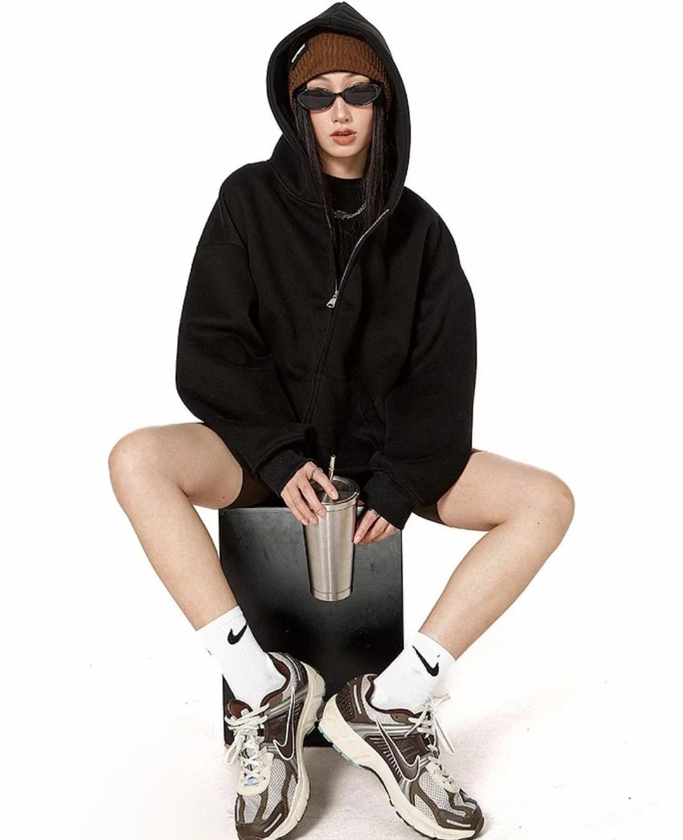 ブラックのオーバーサイズジップパーカーを着てフードをかぶり、ステンレスタンブラーを手に椅子に座るモデルの全身ショット