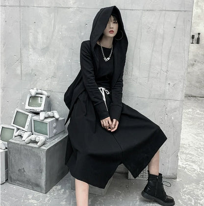 黒の変形フード付きアウターを着たモデル。フードを被り、台に腰掛けて座っている。