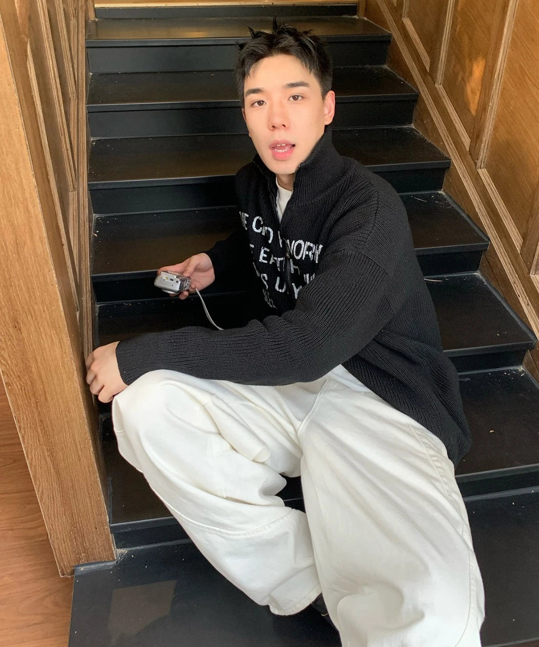 黒の英字ロゴジップアップニットを着た男性モデル。階段でカメラを持つ。