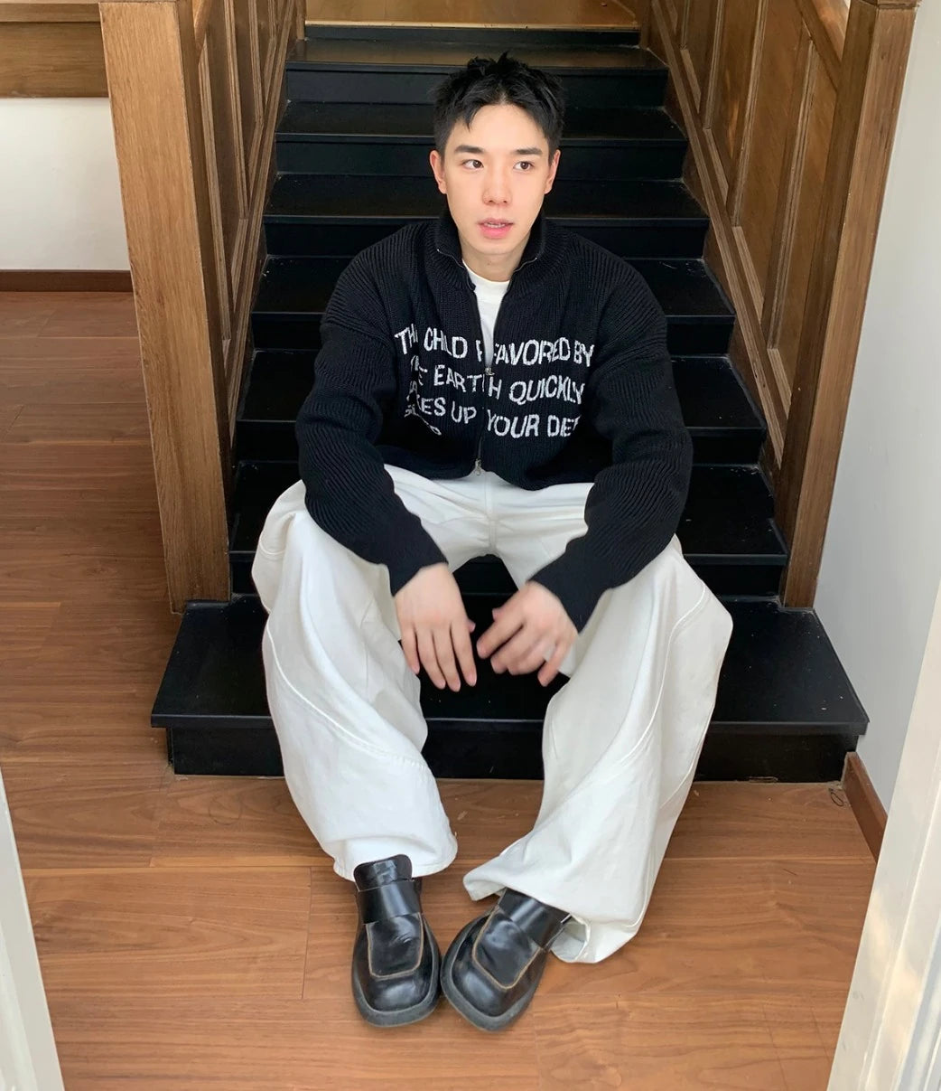 黒の英字ロゴジップアップニットを着た男性モデル。階段に座り、白いワイドパンツ