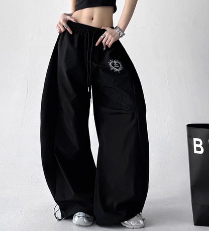 黒いオーバーサイズパンツを着用し、ウエストに手を置いた全身写真。左太もものハート型「Lucky」刺繍ロゴがアクセント。