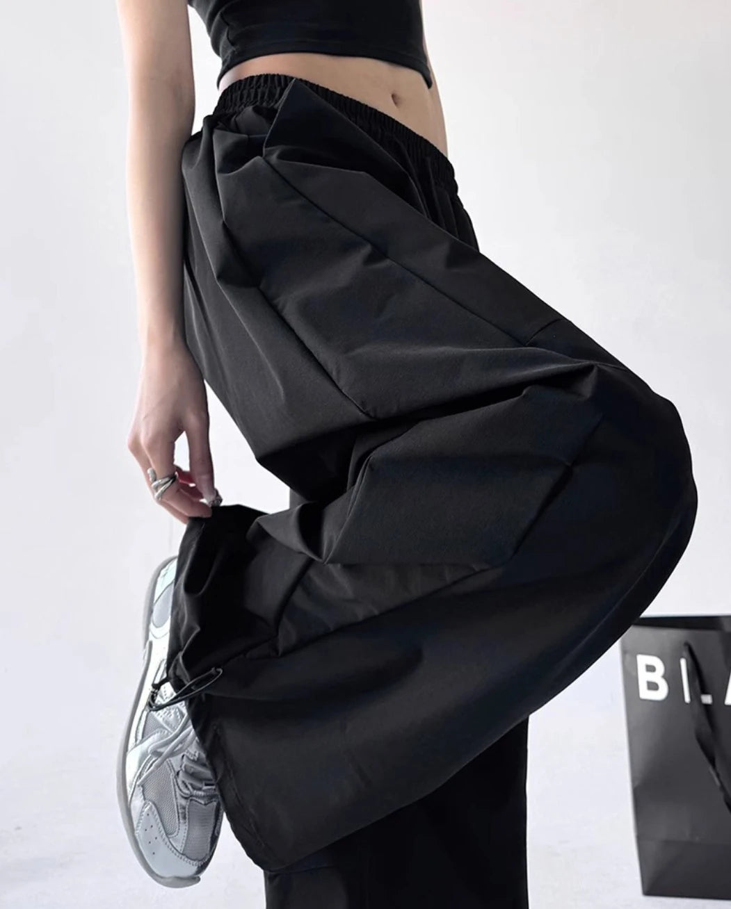 黒いパラシュートパンツを着用し、右足を上げ、ポケットに手を入れたポーズ。左太ももの「Lucky」刺繍ロゴと裾のドローコードが見える。