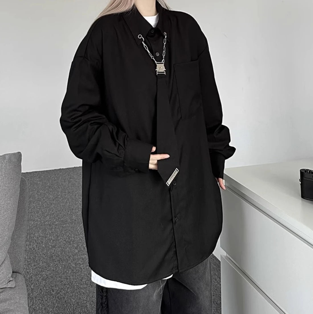 黒のオーバーサイズシャツを着用し、ネクタイ部分に手を添えたポーズ。ユニークなメタルパーツの装飾をアピール。