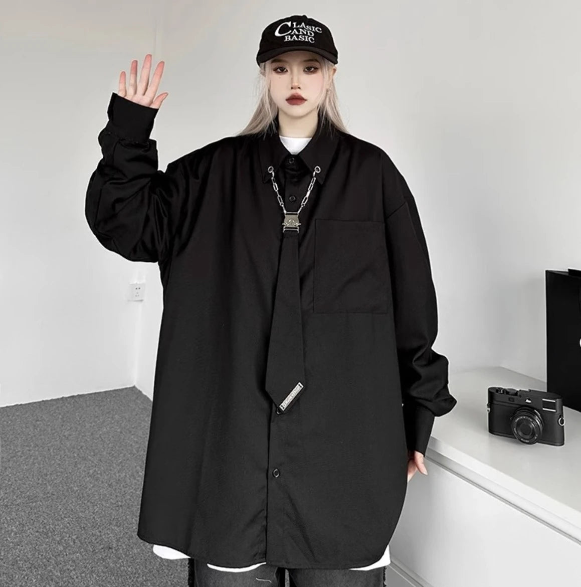 黒のオーバーサイズシャツを着用し、片手を上げたポーズ。黒いキャップを合わせ、ゆったりとしたシルエットを強調。