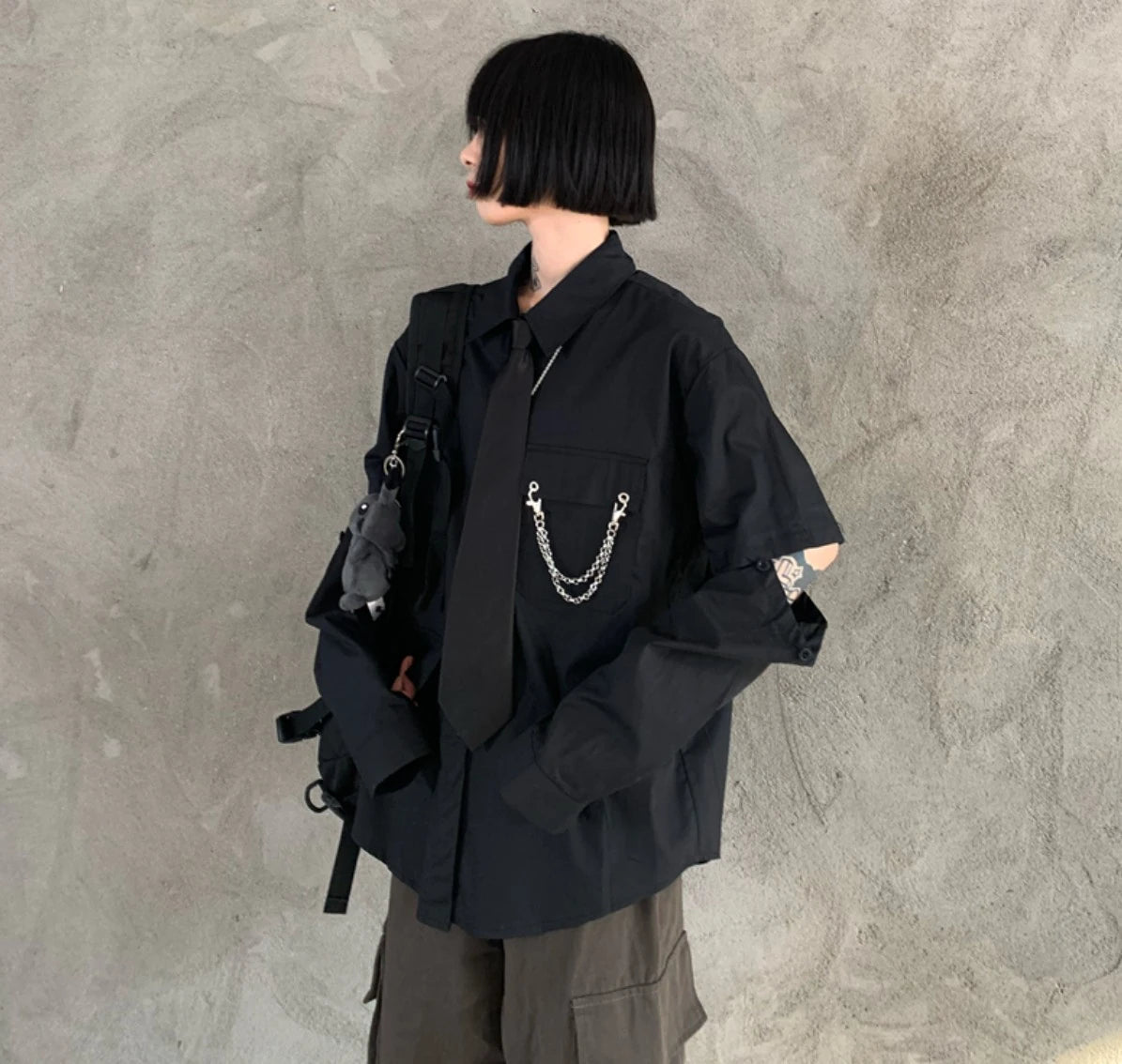 黒のネクタイ付きシャツを着たモデル。横向きで、袖のボタンを外して肌を見せている。