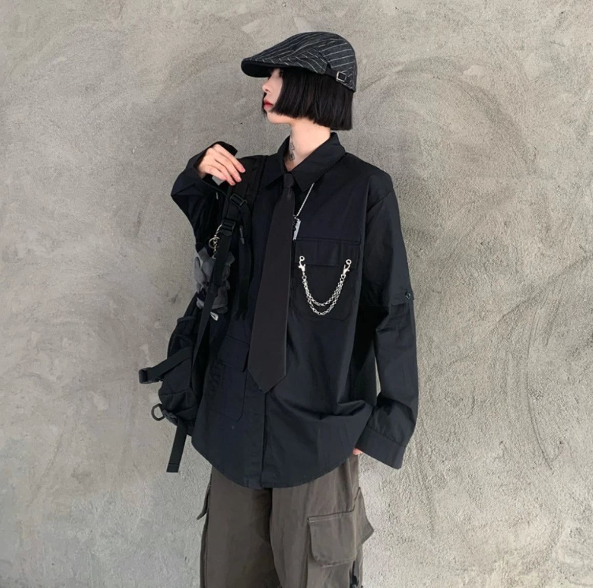 黒のネクタイ付きシャツを着たモデル。ストライプのキャスケットを被り、リュックのストラップを持つ。