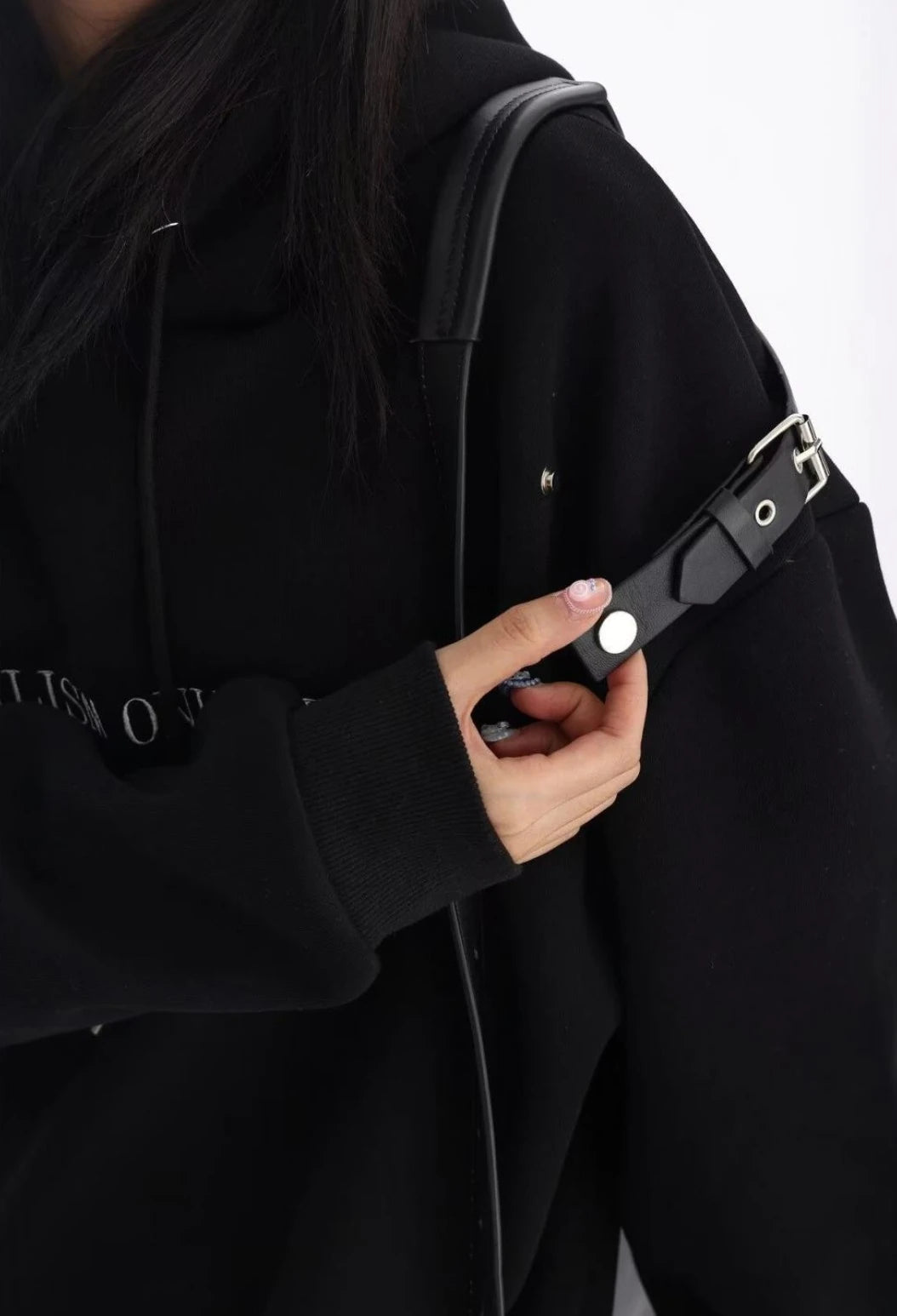 黒のショルダーベルト付きオーバーサイズパーカーの肩ベルト部分を手で押さえたディテールカット。金具デザインがよく分かるショット。