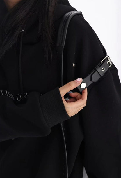 黒のショルダーベルト付きオーバーサイズパーカーの肩ベルト部分を手で押さえたディテールカット。金具デザインがよく分かるショット。
