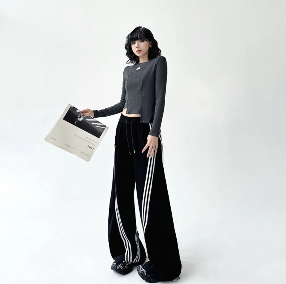 3本ライン入りの黒ワイドパンツを着用し、英字新聞を持ったモデルの全身写真。グレーのトップスと合わせたモードストリートな着こなし。