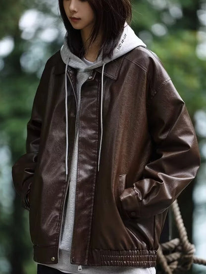 ブラウンのオーバーサイズレザージャケットをフード付きパーカーに重ね着。公園でのクローズアップ。