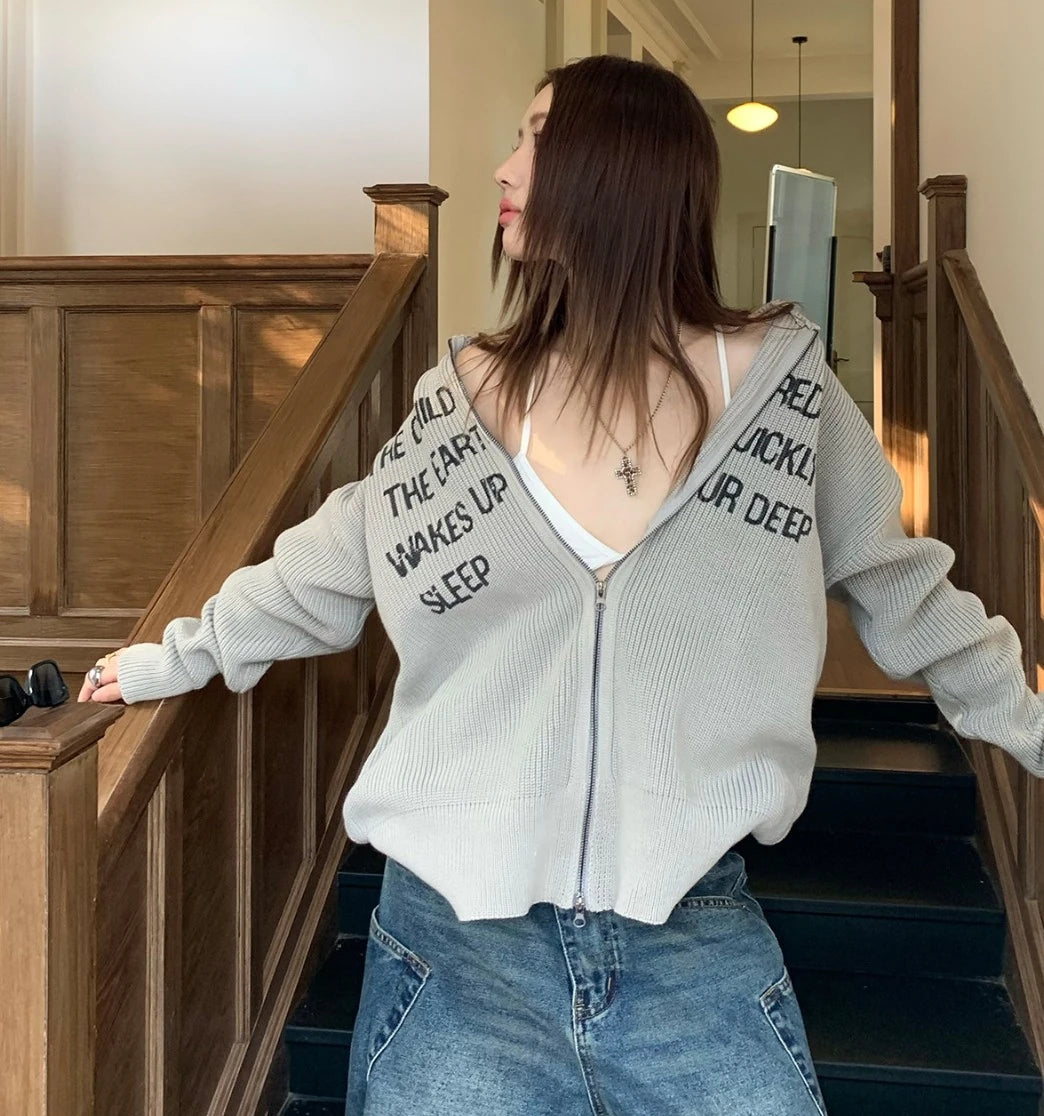 グレーの英字ロゴジップアップニットを着た女性モデル。階段の手すりに寄りかかる。