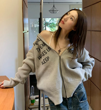 グレーの英字ロゴジップアップニットを肩出しで着た女性モデル。上を見上げる。