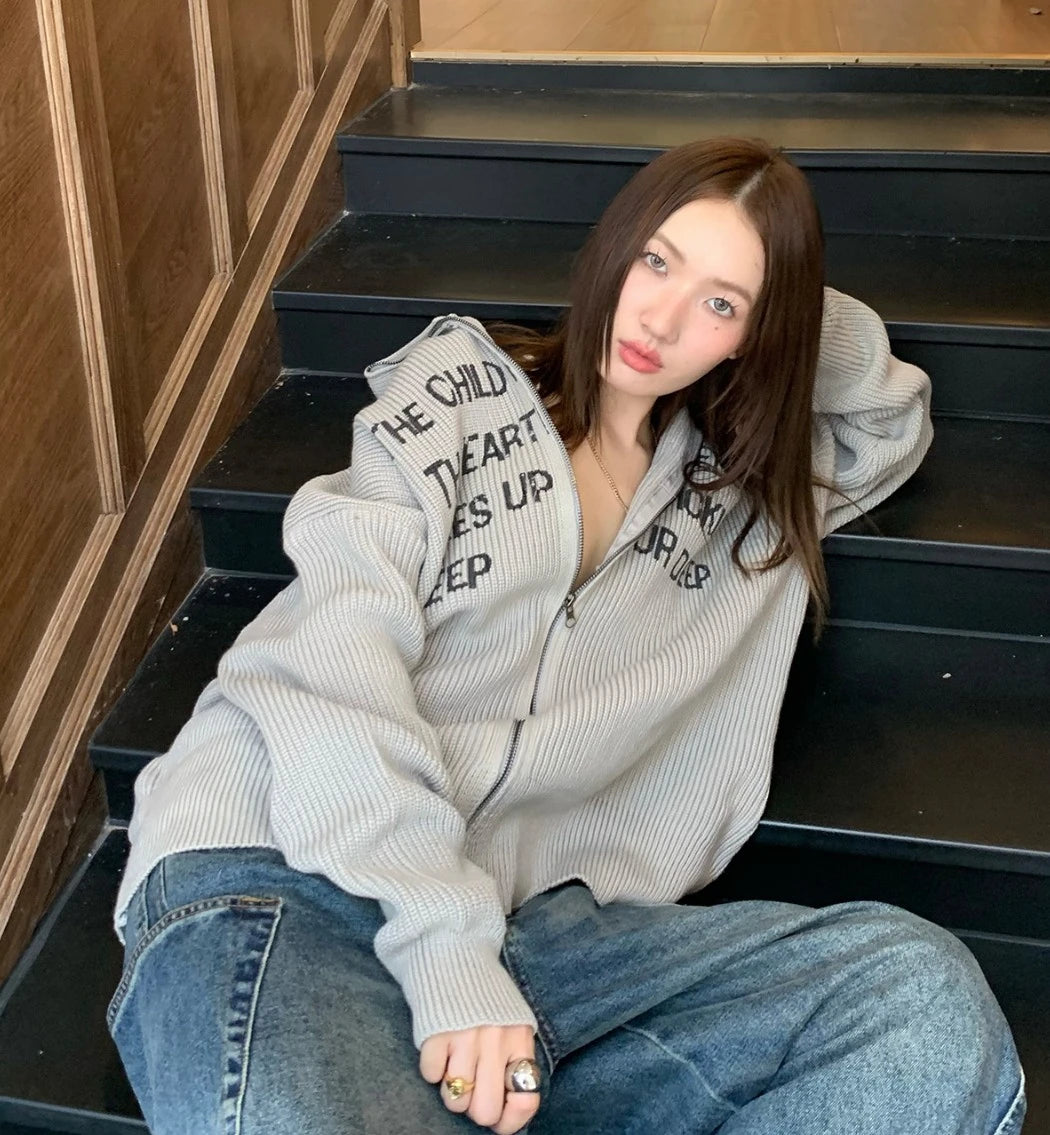 グレーの英字ロゴジップアップニットを着た女性モデル。階段に座り、肘をつく。