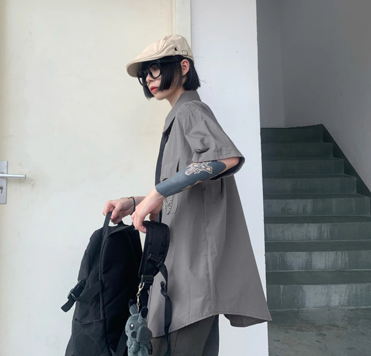グレーのネクタイ付きシャツを半袖風に着たモデル。リュックを手に持ち、階段の前に立つ。