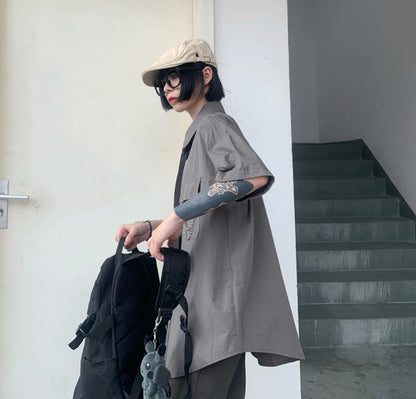 グレーのネクタイ付きシャツを半袖風に着たモデル。リュックを手に持ち、階段の前に立つ。