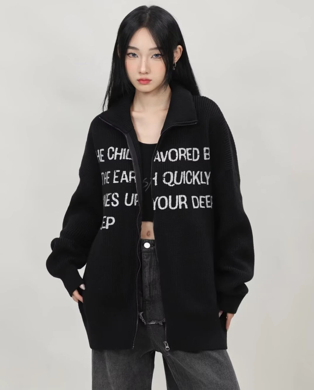 ブラックの英字メッセージジップニットカーディガンをラフに羽織り、ポケットに手を入れたレディースモデルのウエストアップ