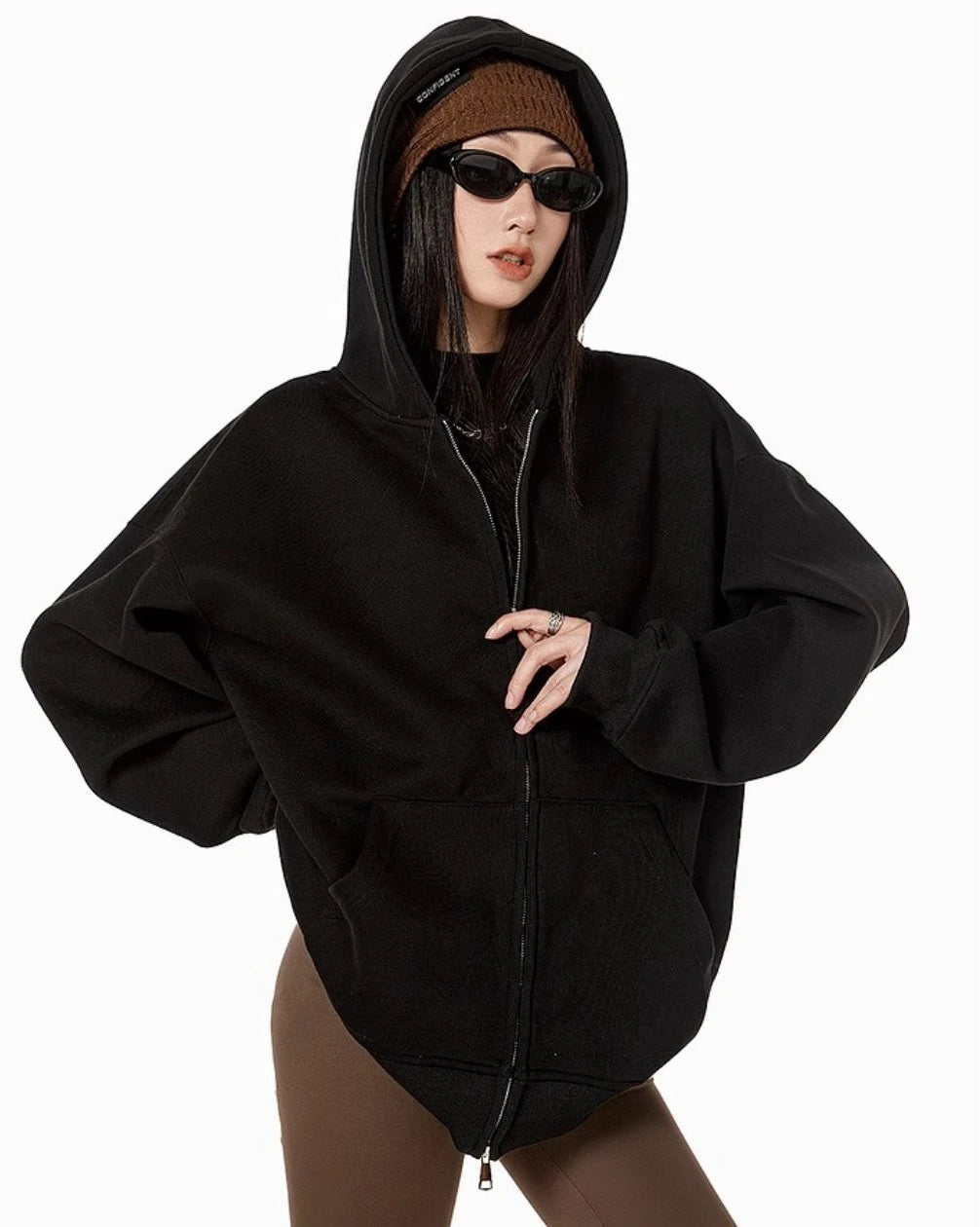 ブラックのオーバーサイズジップパーカーのフードをかぶり、ブラウンのニット帽とサングラスを合わせたモデルの正面ウエストアップ
