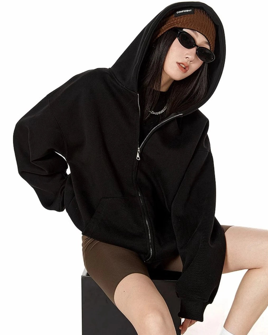 ブラックのオーバーサイズジップパーカーのフードをかぶり、椅子に腰掛けたモデルのリラックスしたポーズ