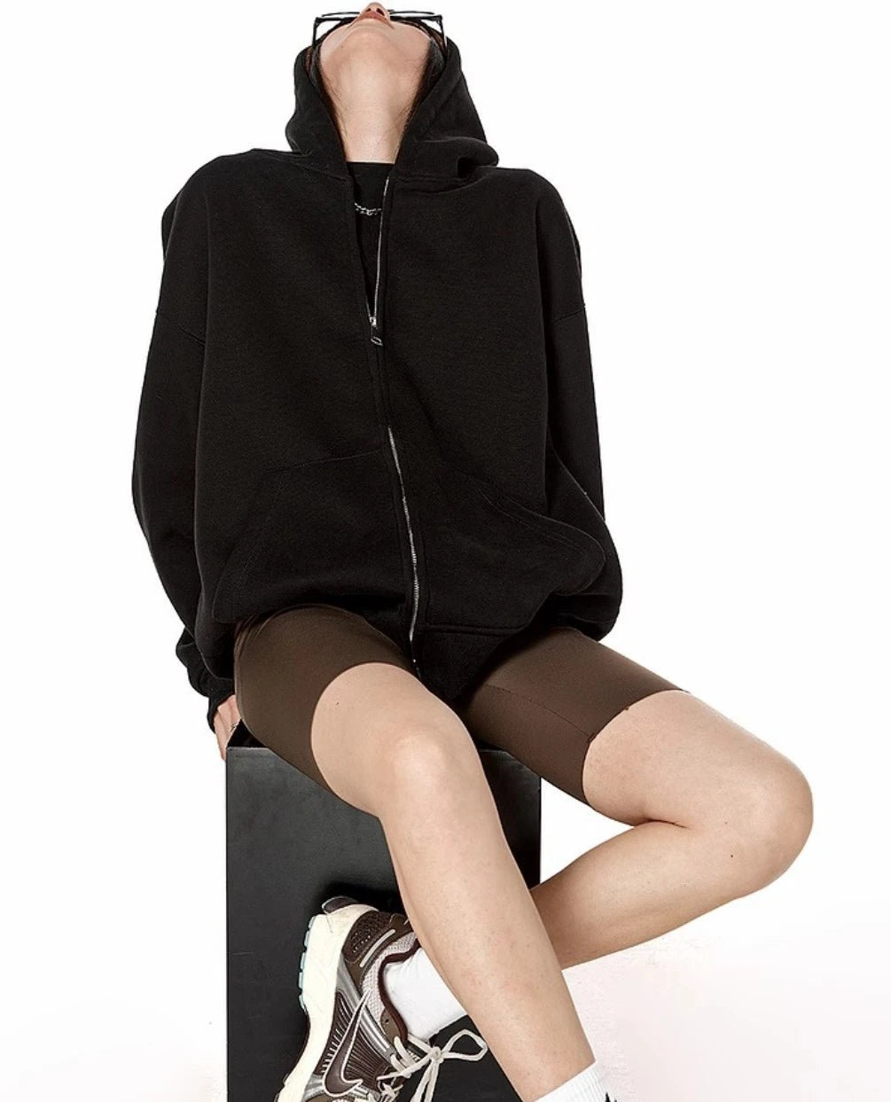 ブラックのオーバーサイズジップパーカーをジップアップし、椅子にもたれて上を向くモデルの座りポーズ