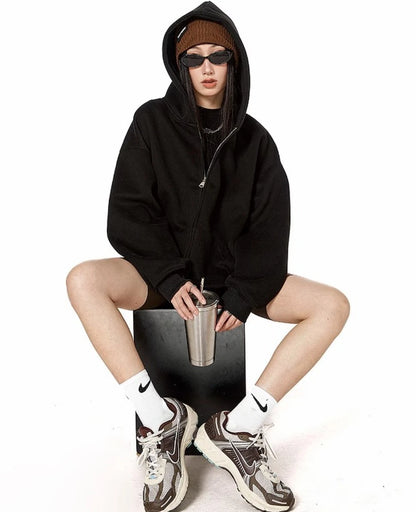 ブラックのオーバーサイズジップパーカーを着てフードをかぶり、ステンレスタンブラーを手に椅子に座るモデルの全身ショット