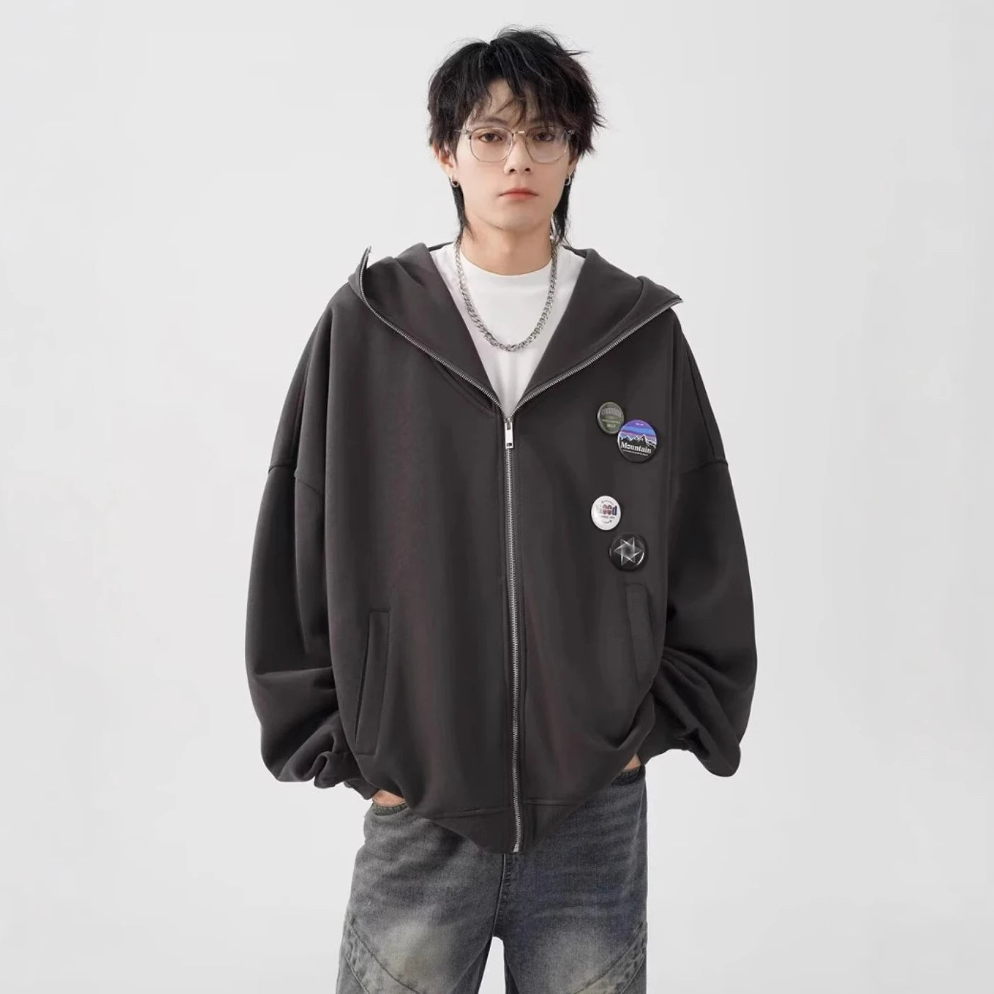 ダークブラウンのオーバーサイズジップパーカーを着用した男性モデルの正面写真。メガネと複数の缶バッジが特徴。