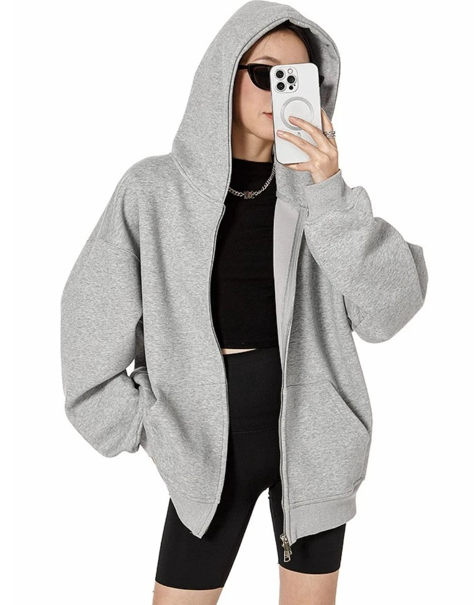 フードをかぶったグレーのオーバーサイズジップパーカーを前開きで羽織り、スマホで顔を隠したモデルの上半身ショット