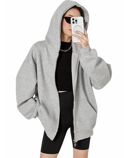 フードをかぶったグレーのオーバーサイズジップパーカーを前開きで羽織り、スマホで顔を隠したモデルの上半身ショット