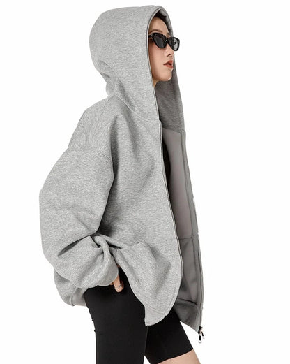 フードをかぶったグレーのオーバーサイズジップパーカーを羽織り、横向きでポケットに手を入れたモデルのサイドショット