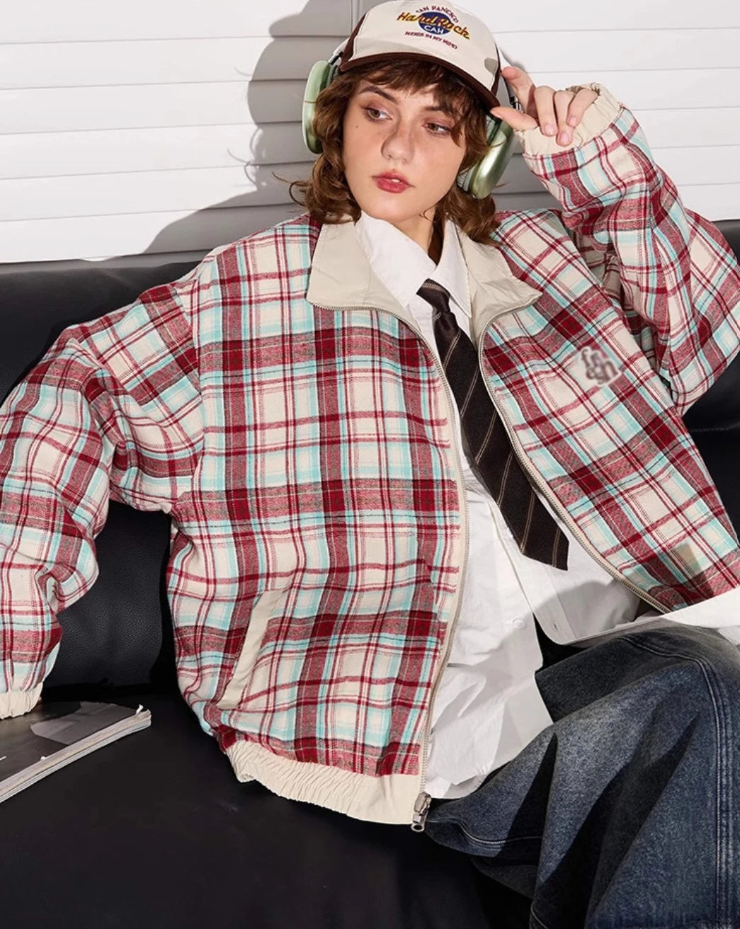 赤チェック柄側のリバーシブルジャケットを着たモデル。ソファに座り、キャップを触る。
