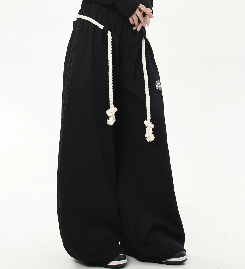 ロングの白ロープ紐が垂れたブラックワイドスウェットパンツを斜め前から撮影した画像。ボリュームのあるワイドシルエットが強調されている。