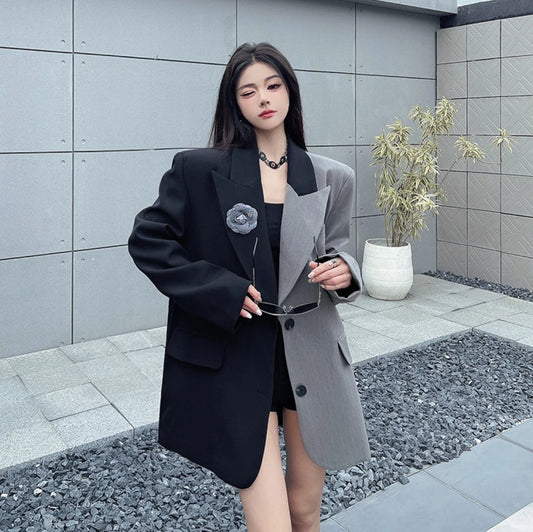 黒とグレーのアシンメトリージャケットを着用した屋外での写真。手にサングラスを持ち、リラックスした表情。