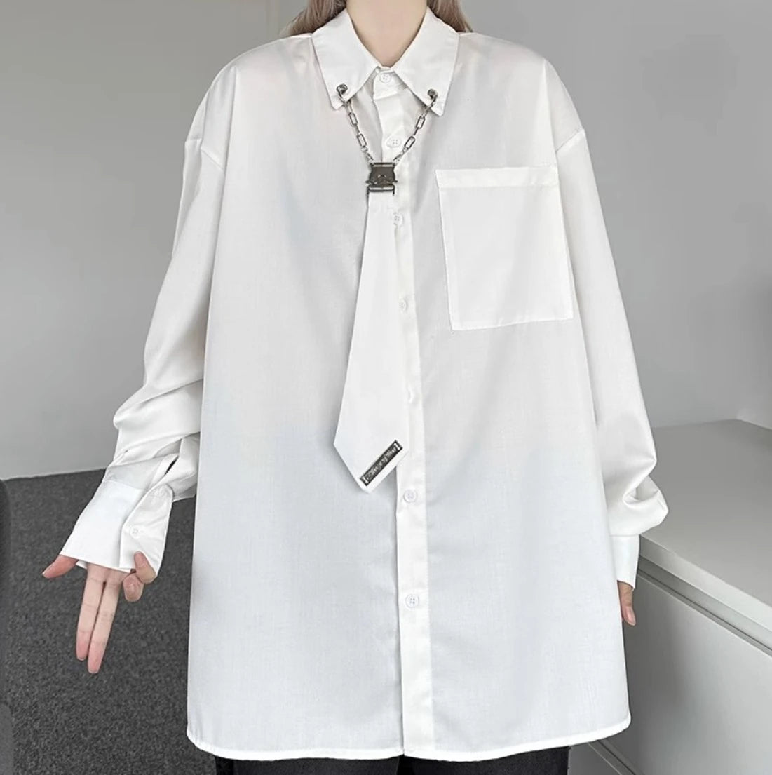 白のオーバーサイズシャツのフロントディテール。首から下のアングルで、襟元のシルバーチェーンとメタルクリップの装飾を強調。