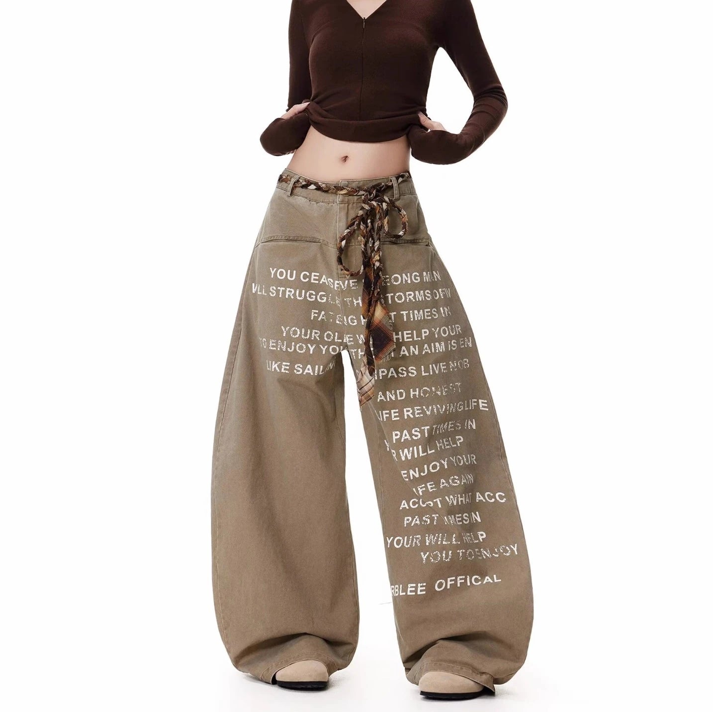 ベージュのワイドレッグパンツを着用し、両手を腰に当てたポーズ。パンツ全体に白の英字プリントが施されている。